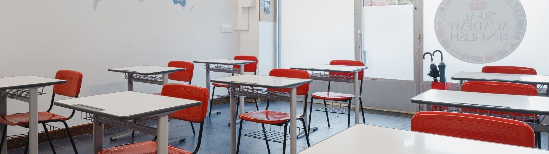 Aula de inglés en The Real Academy Of English con pupitres blancos, sillas rojas y mapa del mundo en la pared